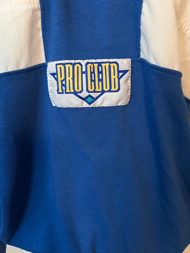 May include: Close-up of a blue and white athletic top. The top features a "PRO CLUB" patch in yellow and blue. The top has a ribbed texture and a classic athletic design.