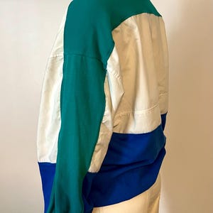 May include: Color-blocked jacket in teal, white, and blue. The jacket features a high collar and long sleeves. The design includes a teal upper section, a white center panel, and a blue lower section. The jacket is made of a lightweight material.