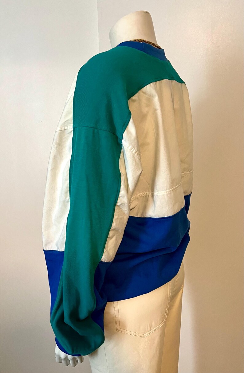May include: Color-blocked jacket in teal, white, and blue. The jacket features a high collar and long sleeves. The design includes a teal upper section, a white center panel, and a blue lower section. The jacket is made of a lightweight material.