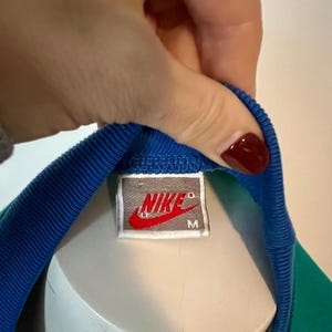 May include: Close-up of a vintage Nike athletic shirt tag. The tag is rectangular with the Nike logo in red and white. The shirt has a blue collar and a green and blue body.