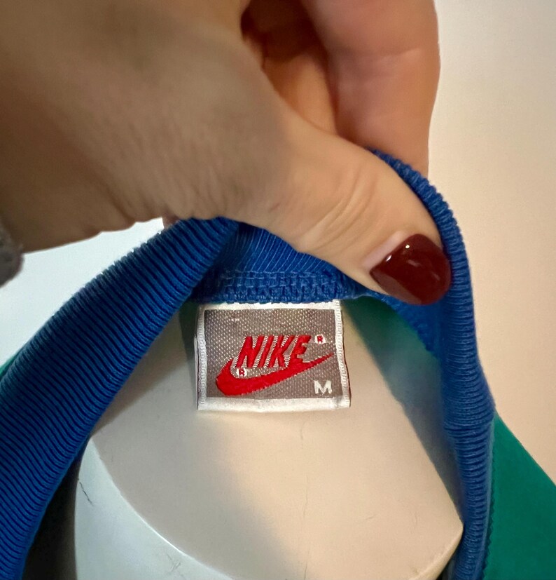 May include: Close-up of a vintage Nike athletic shirt tag. The tag is rectangular with the Nike logo in red and white. The shirt has a blue collar and a green and blue body.