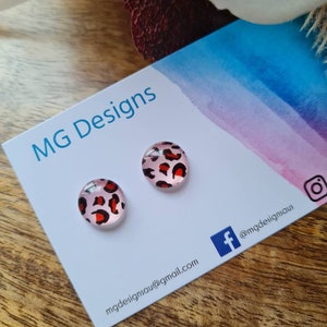 May include: A pair of pink and red leopard print stud earrings on a white card with the text "MG Designs" and contact information. The card also has a blue Facebook icon and a white Instagram icon.