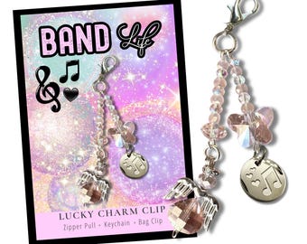 Puede incluir: Un clip de metal plateado con un dije que incluye cuentas facetadas de color rosa claro, un cristal en forma de mariposa, un pequeño dije de ala de ángel y un disco plateado grabado con una nota musical y tres corazones pequeños.