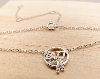 May include: Silver necklace with a circular pendant featuring the letters "BJJ" in a stylized font, surrounded by small clear rhinestones. The pendant also includes a small silver design resembling a belt.