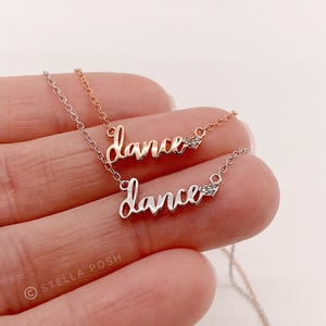 May include: Two delicate necklaces featuring the word dance in cursive script, one in rose gold and one in silver tone. Each necklace includes a small heart-shaped charm accented with tiny clear stones attached to the end of the word.