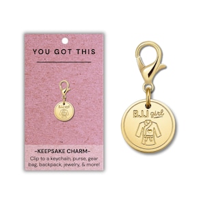 Puede incluir: Un dije circular dorado con cierre de mosquetón, grabado con un kimono de BJJ y el texto BJJ girl. El dije se presenta en una tarjeta rosa con el texto YOU GOT THIS y KEEPSAKE CHARM, junto con una lista de puntos de fijación sugeridos.