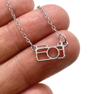May include: A silver-toned necklace featuring a delicate chain and a small, open-frame pendant shaped like a camera. The pendant has a rectangular body with a circular lens cutout and a small square viewfinder detail, suspended from a thin metal link chain.