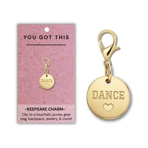 Puede incluir: Un dije circular de tono dorado con un cierre de langosta y la palabra DANCE grabada sobre un pequeño símbolo de corazón. El dije se muestra en una tarjeta rosa con el texto YOU GOT THIS y KEEPSAKE CHARM, junto con instrucciones.