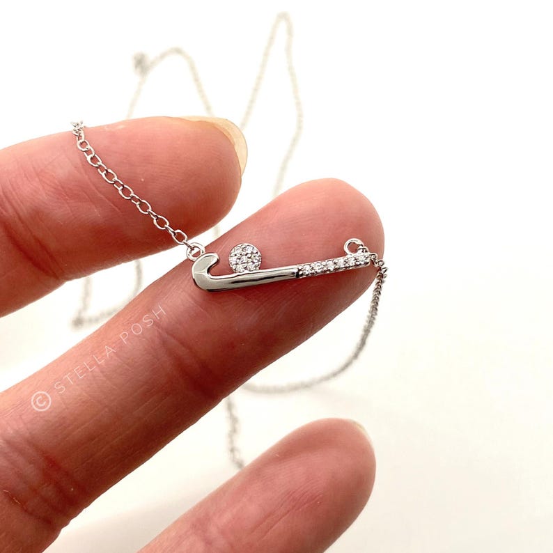 May include: A silver necklace with a hockey-themed pendant. The pendant depicts a hockey stick and ball, both embellished with small, clear stones. The necklace hangs on a fine chain. The brand name "Stella Posh" is visible in the image.
