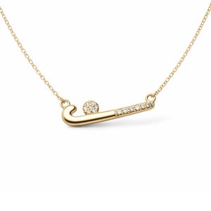 May include: A gold necklace with a field hockey-themed pendant. The pendant depicts a hockey stick and ball, both adorned with small, clear gemstones. The chain is a delicate gold colour. The pendant is approximately 3 cm in length.