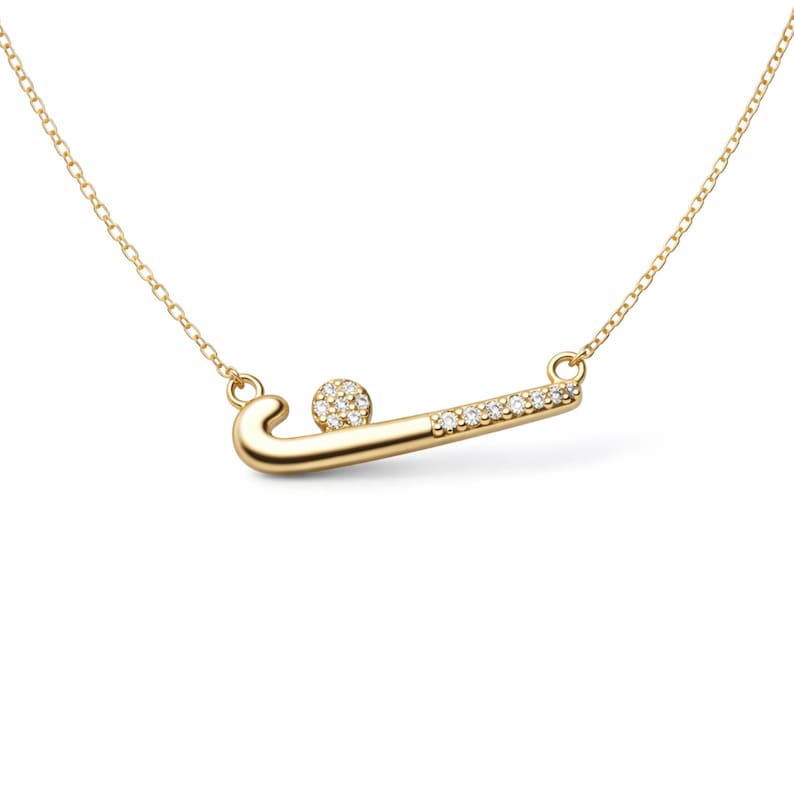 May include: A gold necklace with a field hockey-themed pendant. The pendant depicts a hockey stick and ball, both adorned with small, clear gemstones. The chain is a delicate gold colour. The pendant is approximately 3 cm in length.