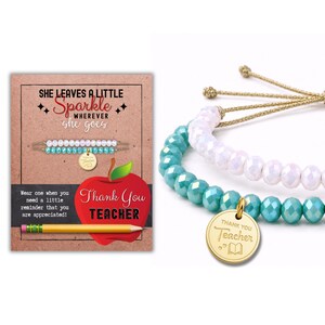 Pode incluir: Duas pulseiras ajustáveis com contas facetadas em branco e turquesa, finalizadas com cordões dourados. Um pingente redondo de metal dourado está preso à pulseira turquesa, gravado com o texto Thank You Teacher e um ícone de livro.