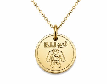 May include: A gold-toned necklace featuring a circular pendant with a polished finish. The pendant is engraved with the text BJJ girl above a graphic illustration of a martial arts gi with a small heart detail on the chest area of the garment.