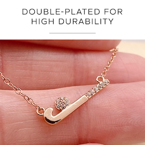 May include: Rose gold necklace with a hockey stick and ball pendant. The hockey stick is decorated with small, clear crystals. The necklace is held by a person. The text "DOUBLE-PLATED FOR HIGH DURABILITY" is at the top.