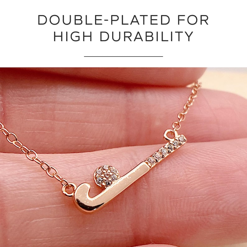 May include: Rose gold necklace with a hockey stick and ball pendant. The hockey stick is decorated with small, clear crystals. The necklace is held by a person. The text "DOUBLE-PLATED FOR HIGH DURABILITY" is at the top.