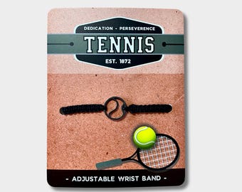 May include: Black adjustable wristband with a tennis ball charm. The packaging features the text "Dedication - Perseverance Tennis Est. 1872" and "Adjustable Wrist Band".