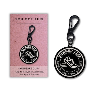 Może przedstawiać: Okrągły czarno-biały brelok z grafiką buta do biegania oraz napisem "RUNNER LIFE" i "keep going - you got this". Brelok posiada czarny plastikowy klips i jest prezentowany na różowej karcie z napisem "YOU GOT THIS".
