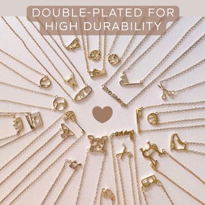 May include: A selection of gold-coloured necklaces with diverse pendant designs, such as sports equipment, scissors, and letters. The image includes the text "DOUBLE-PLATED FOR HIGH DURABILITY". The necklaces are displayed on a white surface.
