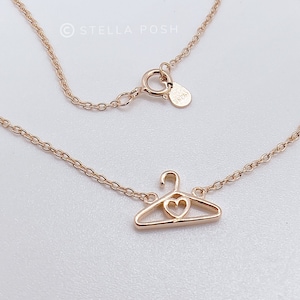 Op de afbeelding: Een gouden ketting met een delicate ketting en een hanger in de vorm van een kleerhanger met een hartvormige uitsparing. De ketting heeft een karabijn sluiting en een klein label met de tekst "925" erop.