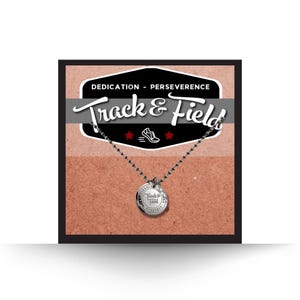May include: A silver necklace with a round pendant that says "Track & Field" on it. The necklace is on a brown background with the words "Dedication - Perseverance Track & Field" in a vintage style font.