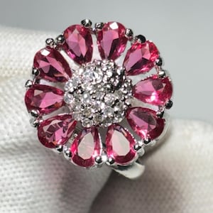 Peut inclure: Une bague en argent avec un motif de fleur rose. La fleur a un centre de petites pierres blanches et des pétales faits de pierres roses plus grandes.