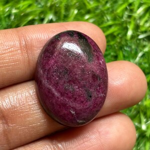 May include: An oval-shaped cabochon gemstone featuring a deep magenta and dark purple colour palette with visible black inclusions throughout the polished surface, showcasing a smooth, rounded top and a reflective, glossy finish.