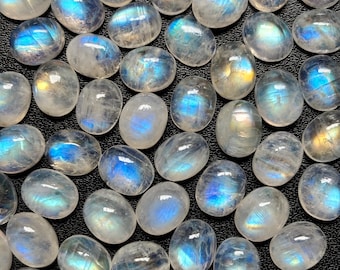 May include: A collection of oval-shaped moonstone cabochons. The stones display a translucent, milky white base with iridescent blue and gold flashes. The gemstones are arranged closely together, filling the frame.