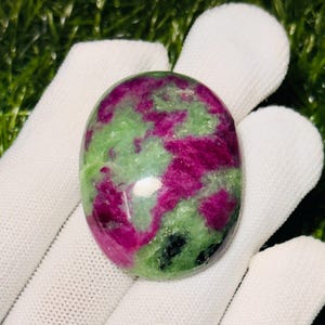 Könnte beinhalten: Ein ovaler, polierter Edelstein-Cabochon mit einem gesprenkelten Muster aus leuchtend magentafarbenen und hellgrünen Mineralieneinschlüssen, wobei kleine schwarze Punkte auf der glatten, reflektierenden Oberfläche zu sehen sind.