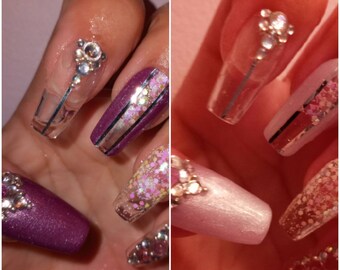 Op de afbeelding: Close-up van twee handen met lange, heldere acrylnagels versierd met glitter, strass en zilveren strepen. De nagels hebben een kistvorm en zijn geverfd in tinten roze en paars.