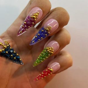 May include: A hand showing long, pointed nails with a pale pink base. Each nail has a different colour of textured gel, including red, green, blue, and purple, decorated with gold beads. The nails are a vibrant and artistic display.