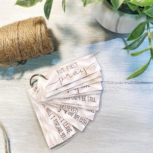 May include: A set of small cards with inspirational quotes on a pink and white chevron background. The cards are attached to a ring and have the words "But First Pray" on the top card.