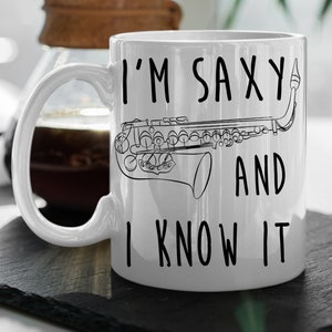 Puede incluir: Taza de cerámica blanca con el texto "I'M SAXY AND I KNOW IT" en negro. Un dibujo lineal de un saxofón aparece en la taza. La taza tiene un asa blanca y está sobre una superficie negra.