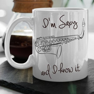 Puede incluir: Taza de cerámica blanca con un dibujo lineal negro de un saxofón y el texto "I'm Saxy and I know it." La taza tiene un asa grande y está destinada a bebidas. Una cafetera es visible en el fondo.