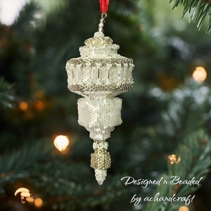 Puede incluir: Un adorno navideño blanco de cuentas con forma de lámpara con una parte superior dorada y una cinta roja que cuelga de la parte superior. El adorno está colgado de un árbol de Navidad verde. El texto "Designed & Beaded by achandercraft" está en la parte inferior de la imagen.