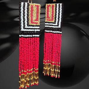 May include: A pair of handmade beaded earrings. The earrings feature a rectangular top with a geometric pattern in black, white, and gold. Below, long strands of red beads cascade down, with gold beads at the bottom. The earrings hang from gold-colored hooks.