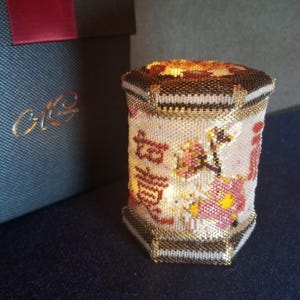 Pode incluir: Uma lanterna decorativa hexagonal com contas, caracteres chineses e desenhos florais. A lanterna é principalmente branca com detalhes dourados, castanhos e vermelhos. Uma caixa azul escura com letras douradas está no fundo.