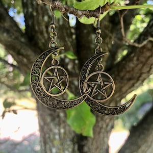 May include: Gold tone crescent moon earrings with a pentagram charm hanging from each moon. The earrings are hanging from a tree branch.