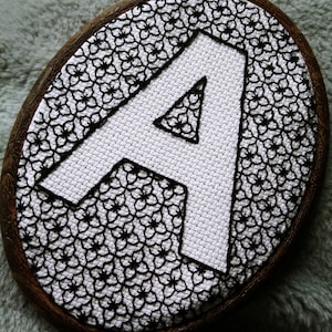 Könnte beinhalten: Ein weißer Stickrahmen mit einem schwarzen gestickten Buchstaben A in der Mitte. Der Buchstabe A ist von einem schwarzen Blumenmuster umgeben.