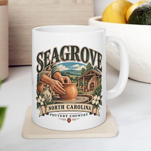 Könnte beinhalten: Ein weißer Keramikbecher mit einer farbenfrohen Illustration eines Töpfers, der Ton auf einer Scheibe formt. Der Text lautet SEAGROVE NORTH CAROLINA POTTERY COUNTRY. Das Design zeigt einen Brennofen, Bäume, Blumen und ein kleines Töpfer-Symbol.