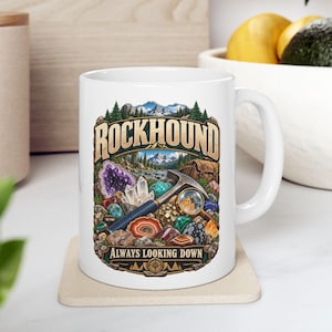 Könnte beinhalten: Ein weißer Keramikbecher mit einer farbenfrohen Grafik einer geologischen Szene und dem Text ROCKHOUND sowie ALWAYS LOOKING DOWN. Die Abbildung zeigt einen Geologenhammer, eine Lupe, verschiedene Kristalle und Steine vor einer Berglandschaft.