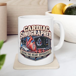 Peut inclure: Un mug en céramique blanc orné d'un graphique représentant un cœur humain, un échographe et le texte "CARDIAC SONOGRAPHER SEEING THE HEART DIFFERENTLY" avec les mots "IMAGING", "ASSESSING", "GUIDING" et "CARING" sur une bannière.