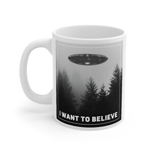 Peut inclure: Une tasse en c&eacute;ramique blanche avec une image en noir et blanc d'un OVNI volant au-dessus d'une for&ecirc;t. Le texte "I WANT TO BELIEVE" est imprim&eacute; en noir au bas de la tasse.