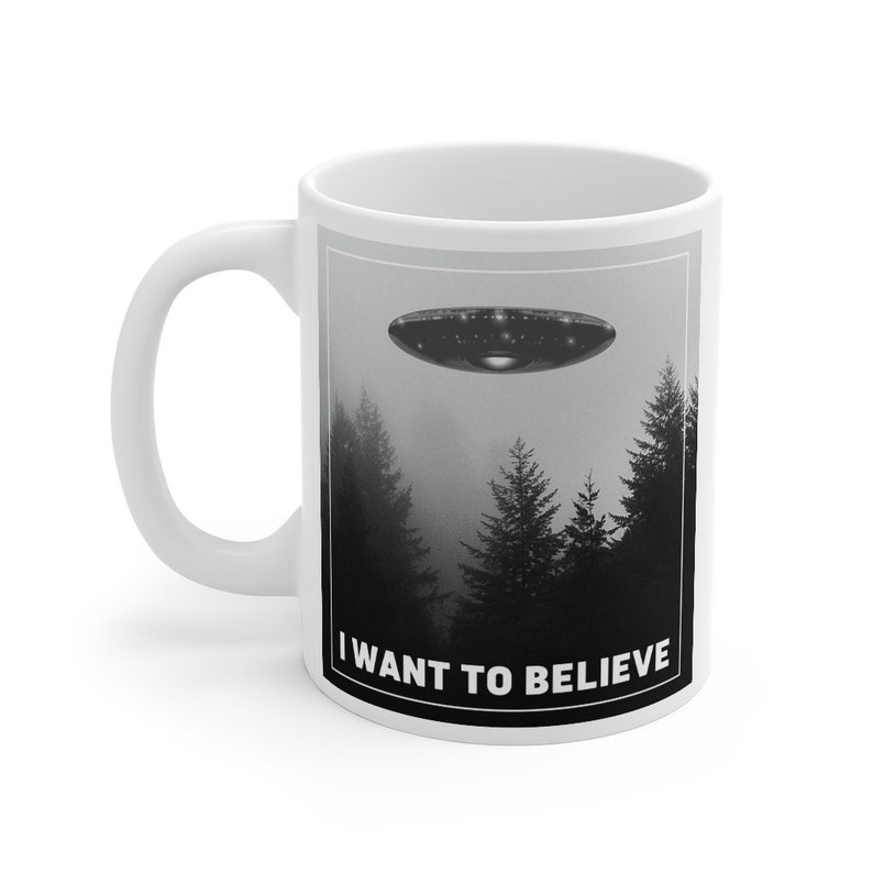 Peut inclure: Une tasse en c&eacute;ramique blanche avec une image en noir et blanc d'un OVNI volant au-dessus d'une for&ecirc;t. Le texte "I WANT TO BELIEVE" est imprim&eacute; en noir au bas de la tasse.