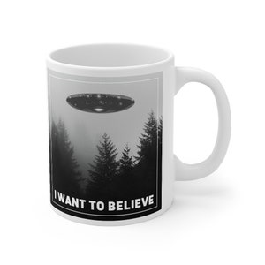 Peut inclure: Une tasse en c&eacute;ramique blanche avec une image en noir et blanc d'un OVNI volant au-dessus d'une for&ecirc;t. Le texte "I WANT TO BELIEVE" est imprim&eacute; sur la tasse.