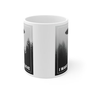 Peut inclure: Une tasse en c&eacute;ramique blanche avec une image en noir et blanc d'un OVNI au-dessus d'une for&ecirc;t. Le texte "I WANT TO BELIEVE" est sur le c&ocirc;t&eacute; droit de la tasse et le texte "BELIEVE" est sur le c&ocirc;t&eacute; gauche de la tasse.