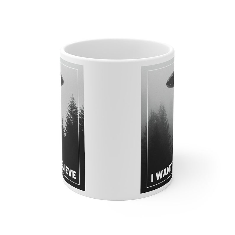 Peut inclure: Une tasse en c&eacute;ramique blanche avec une image en noir et blanc d'un OVNI au-dessus d'une for&ecirc;t. Le texte "I WANT TO BELIEVE" est sur le c&ocirc;t&eacute; droit de la tasse et le texte "BELIEVE" est sur le c&ocirc;t&eacute; gauche de la tasse.