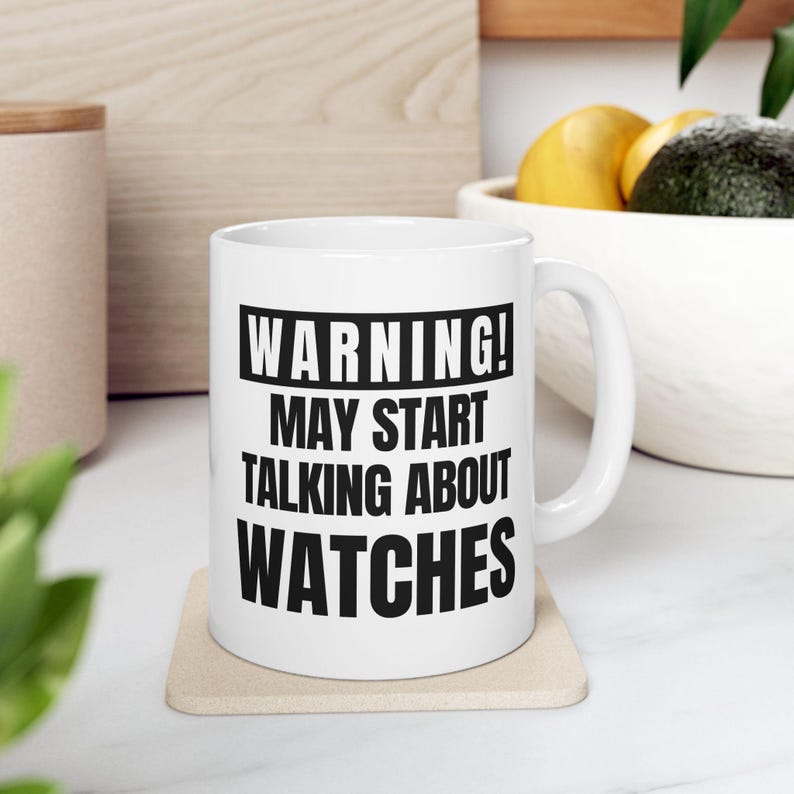 May include: White ceramic mug with the text "WARNING! MAY START TALKING ABOUT WATCHES" in black. The mug is on a beige coaster. The background includes a wooden cutting board, a bowl with fruit, and a wooden container.