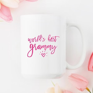 May include: White ceramic mug with the text "world's best grammy" in pink script. A small heart is below the word "grammy". The mug has a large handle and is surrounded by pink flowers.