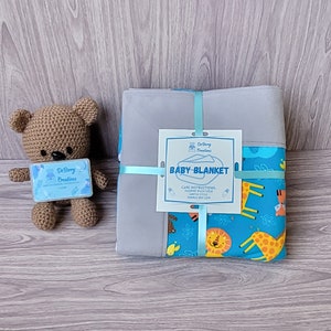 May include: A gray baby blanket with a blue animal print border, tied with a light blue ribbon. A crocheted brown teddy bear with a tag that reads "De Barry Creations" is next to the blanket.