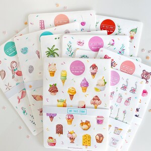 Può includere: Un set di 10 fogli di adesivi con vari temi, tra cui sirene, gelati, creature del bosco e motivi floreali. Ogni foglio ha uno sfondo rosa con bordi bianchi e illustrazioni colorate. I fogli di adesivi sono etichettati con il tema e il nome "Art Tales Studio".